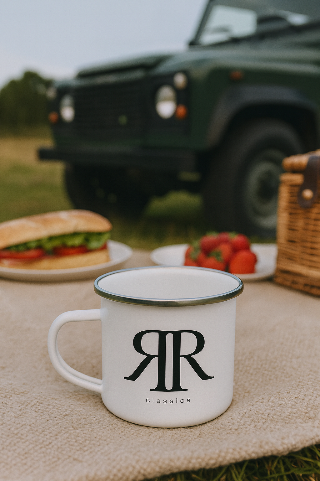 White enamel mug with 'RR' logo on a picnic blanket with a car and food in the background